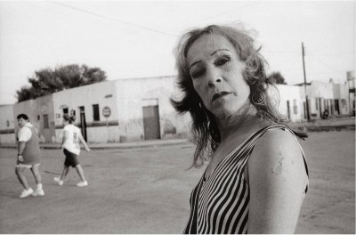 A black-and-white photo of an older woman in heavy makeup looks at the camera in a barrio. 