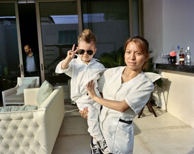 A nanny holds a tolder who has on big sunglasses, a faux hawk, and does a piece sign. 