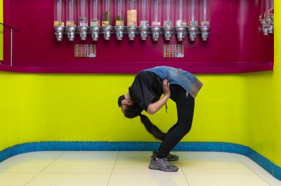 A worker wearing an apron and face masks bends backward in a dancerly pose in front of a yellow-and-magenta painted wall. ON the wall are tubs of cereal. 