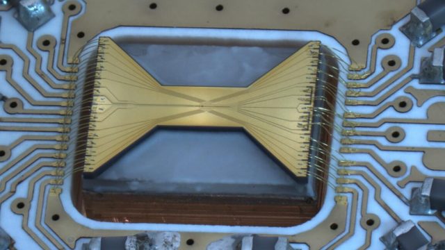 Quantum-teleportation-used-to-distribute-a-calculation.jpeg Image of a gold, bow-tie shaped object, with lots of wires extending out from each end.