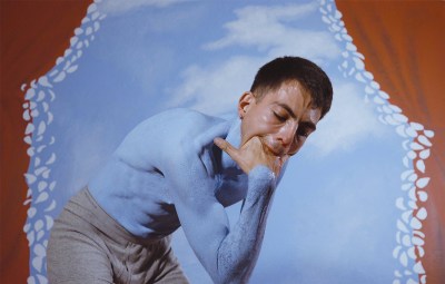 A film still showing a man with a sky blue–painted torso sticks his hand into his mouth in front of a sky background with red curtains on the edge.
