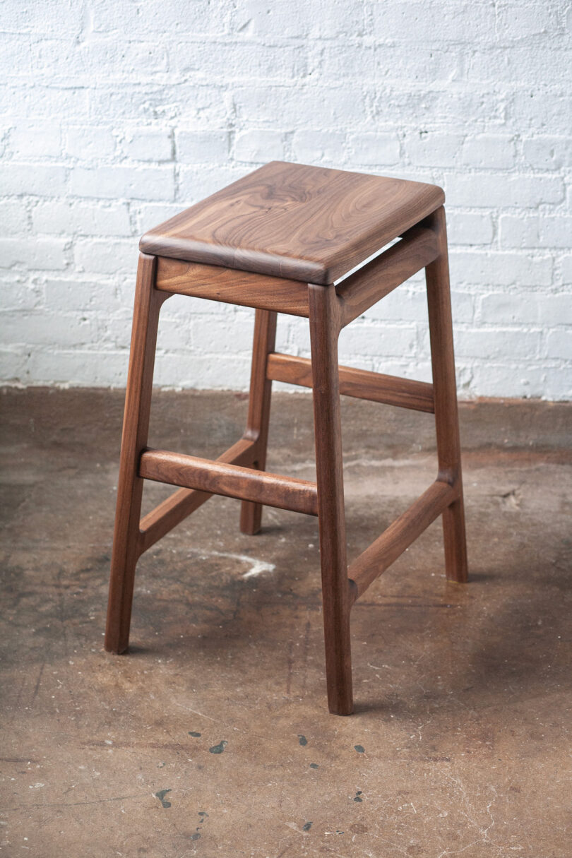 A wooden stool with a square seat and four angled legs, placed on a concrete floor against a white brick wall