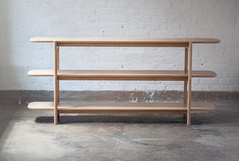 A wooden three-tier shelf stands against a white brick wall on a concrete floor