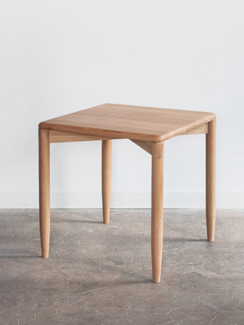 A wooden table with four tapered legs stands on a concrete floor against a plain white wall