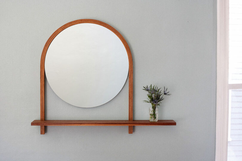 Round mirror with a wooden frame attached to a shelf. A small vase with greenery sits on the shelf against a light-colored wall.