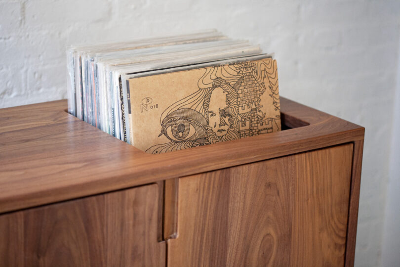 A wooden cabinet with vinyl records, featuring an album with intricate black and white artwork on top