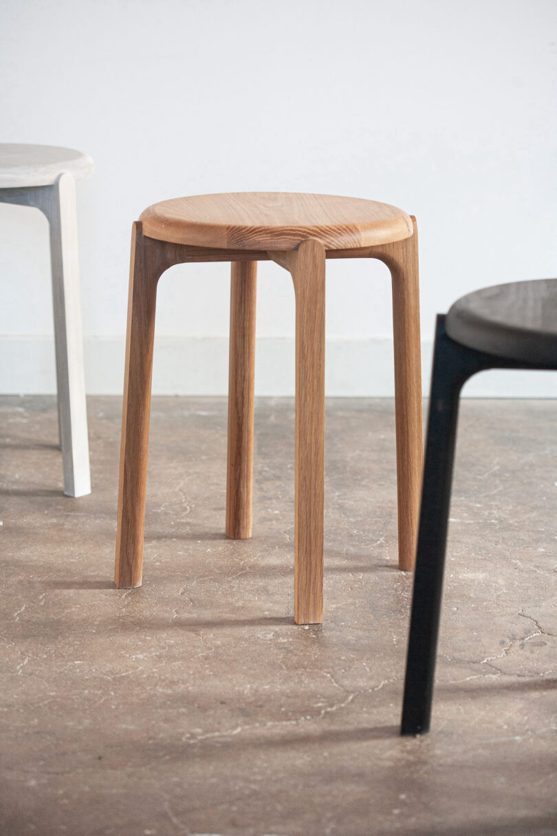 A wooden stool with three legs is centered between a white stool and a black stool on a concrete floor