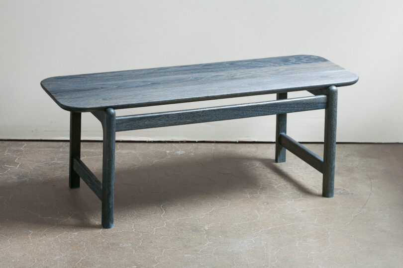 A wooden bench with a simple rectangular seat and four legs stands on a stone floor against a light-colored wall