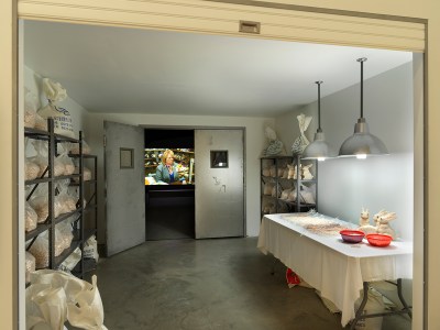 An installation resembling a factory with a worktable. An open doorway shows a video of a woman at a similar table.