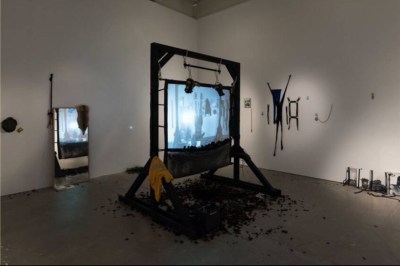 An installation featuring a hanging screen above various detritus. An image of a person hanging upside down is projected onto the screen.