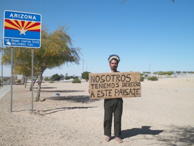 A man holding a sign by the Arizona state border saying 'NOSOTROS TENEMOS DERECHO A ESTE PAISAJE.'