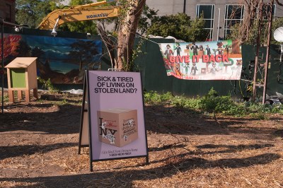 An area with woodchips with a clapboard sign that reads 'SICK & TIRED OF LIVING ON STOLEN LAND?'