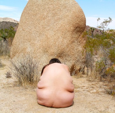 A nude woman seen from behind posed before a large boulder.