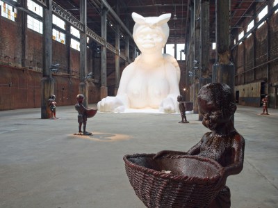 A giant sphinx made out of sugar with large breasts and a bandana. The sphinx is situated in a disused factory, with several small sculptures of children holding out baskets around it.