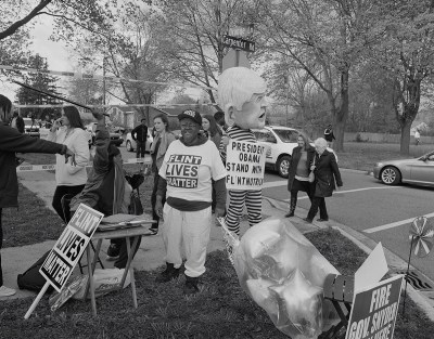 A black-and-white photograph of a protest with one person wearing a shirt that says 'FLINT LIVES MATTER.'
