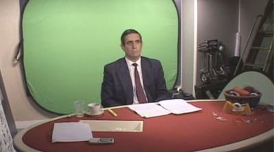 A man in a suit sitting at a desk in front of a green screen.