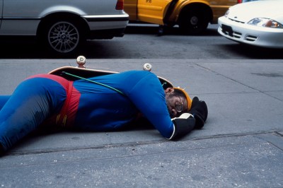 A Black man in a Superman costume with a skateboard strapped to his back collapsed on a sidewalk.