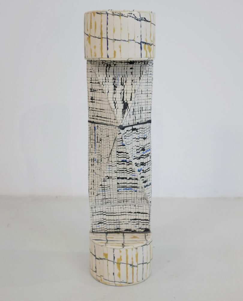 Tall ceramic sculpture with a cylindrical shape, featuring a cracked, grid-like pattern in black and beige on a white background.