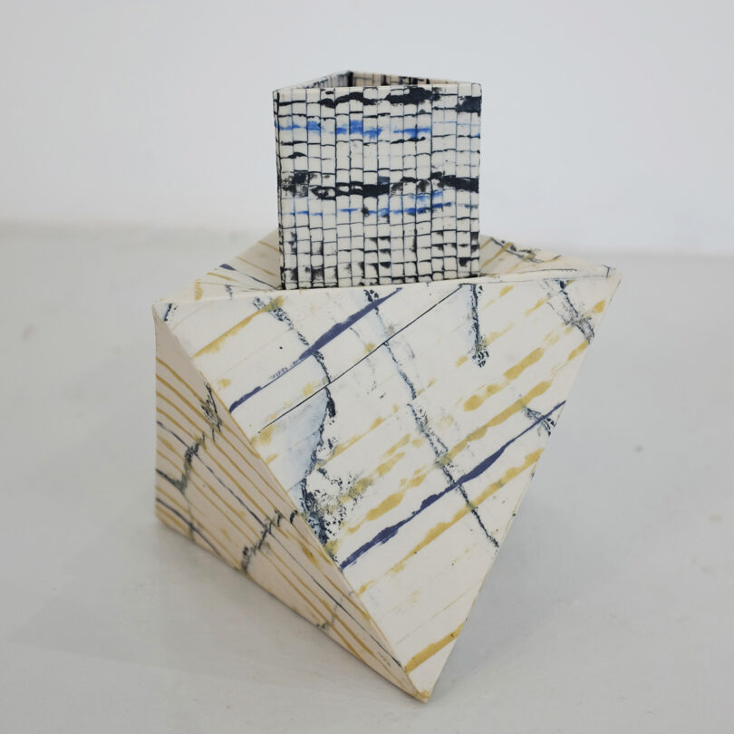 Geometric sculpture featuring a pyramid and a cube. The pyramid has diagonal stripes, while the cube has a grid pattern. Colors include white, cream, blue, and black.