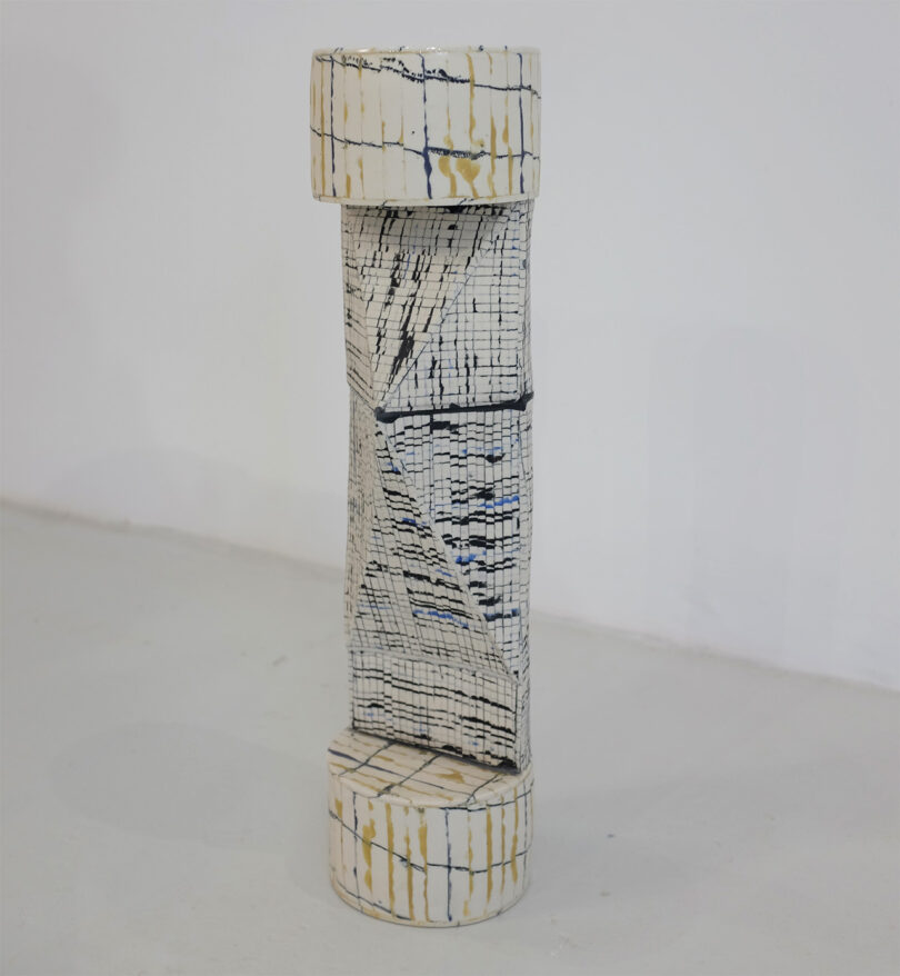 A tall, cylindrical ceramic sculpture with a cracked pattern and beige and blue lines stands against a plain background.