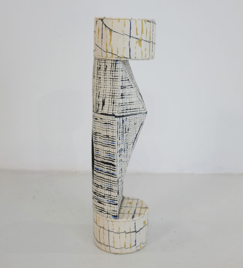 Abstract ceramic sculpture with geometric shapes, featuring vertical lines and grids in black and beige on a white surface, displayed against a plain background.