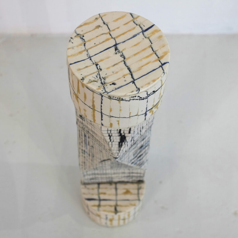 Tall, cylindrical sculpture with beige and dark line patterns placed on a light surface.