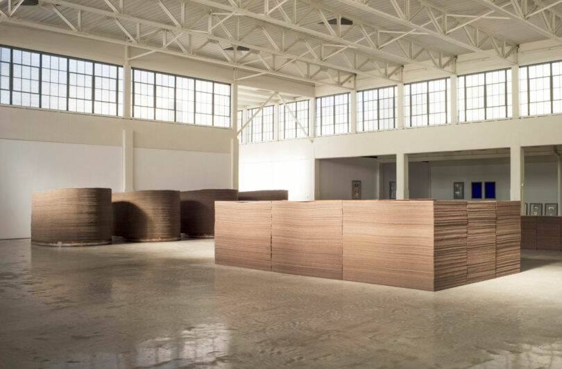 Stacks of large cardboard sheets are arranged in a spacious, well-lit warehouse with high ceilings and large windows.