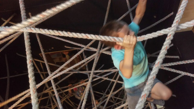 Child climbing through a web of ropes in an indoor activity area.
