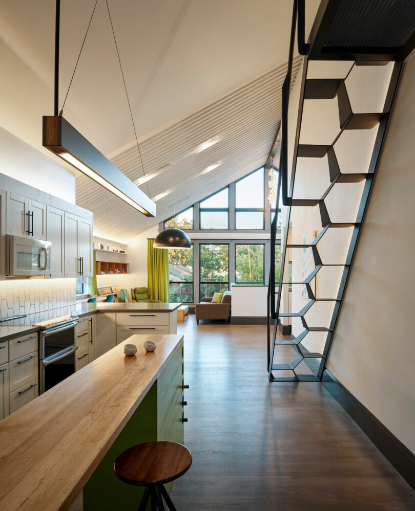 Modern loft apartment with sloped ceiling. Features a kitchen island, wooden flooring, hexagonal staircase, and large windows overlooking greenery. Minimalist decor with a couch and dining area.