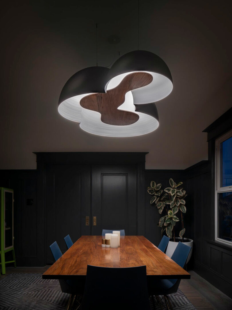 Modern dining room with a wooden table, blue chairs, a leafy plant, and a large, artistic ceiling light fixture with a wooden accent.