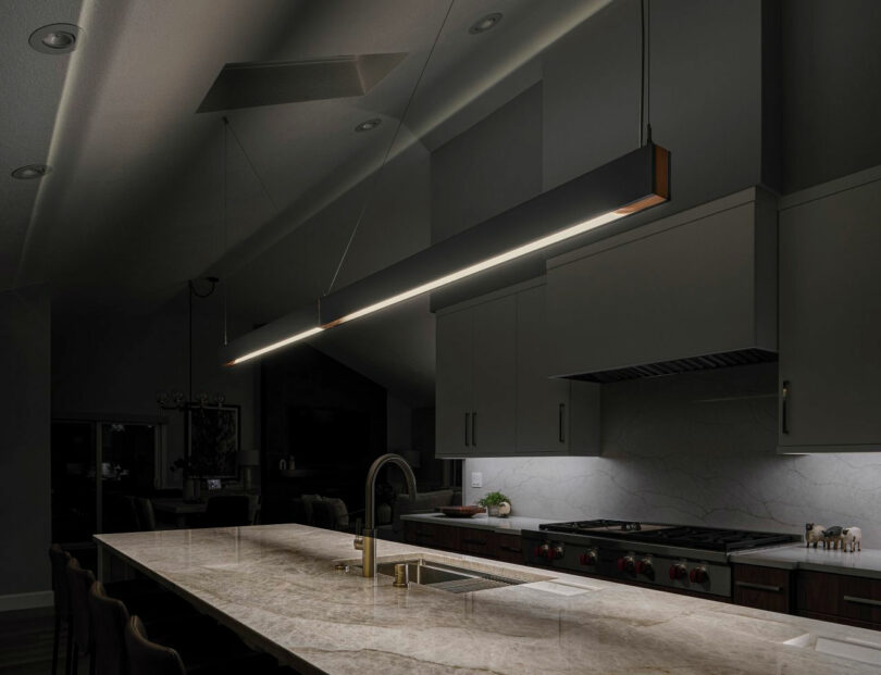 Modern kitchen with a long marble island, stainless steel sink, overhead pendant light, and integrated appliances in a dimly lit setting.