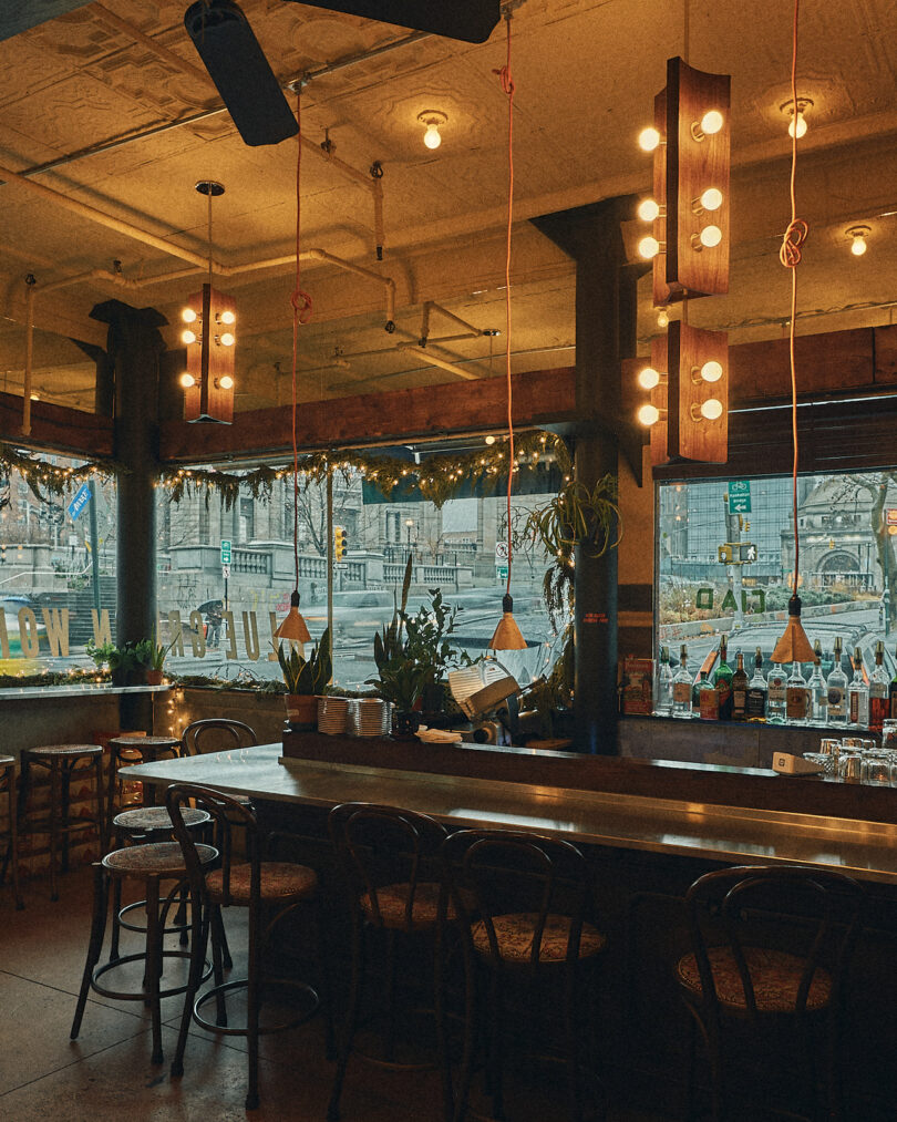 A cozy bar interior with bar stools and a counter, decorated with hanging lights and plants. Large windows provide a view of the street outside