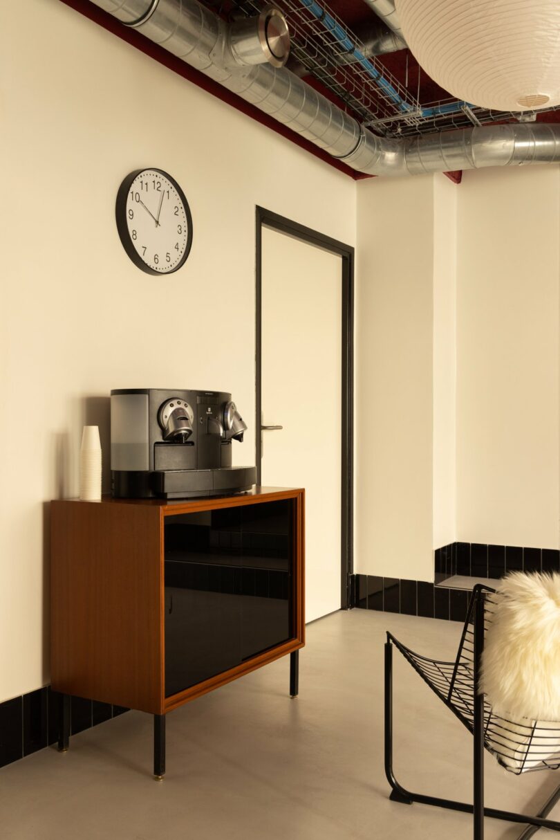Office space with a clock on the wall showing 3:55, a coffee machine on a wooden cabinet, and a black wire chair with a fluffy cushion. Pipes are visible on the ceiling.