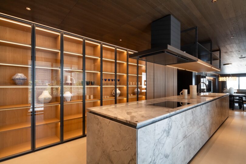 Modern dining area with a round table, dark chairs, and a decorative light fixture. Warm lighting highlights the wood ceiling and wall panels.