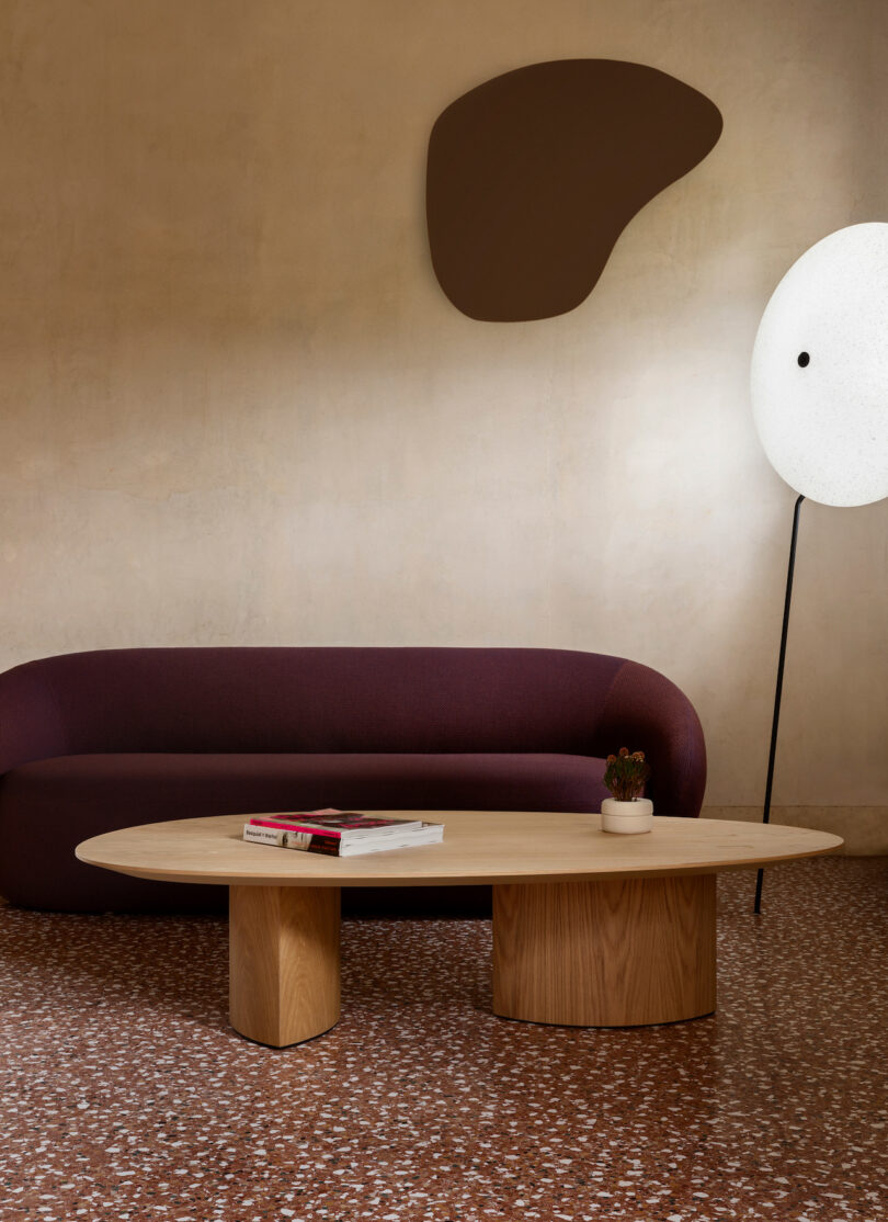 A modern living room with a maroon couch, oval wooden coffee table, abstract wall art, and a round floor lamp. Carpet features a terrazzo pattern