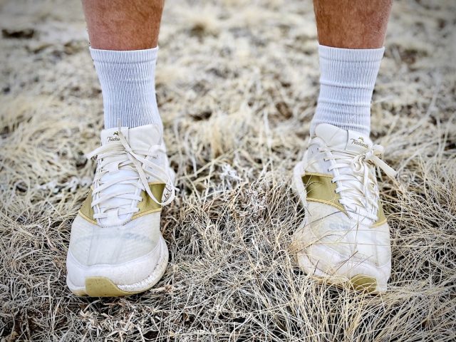 1742329084_Tracksmith-Eliot-Racer-Review-GearJunkie.jpeg A pair of Tracksmith Eliot Racer shoes worn with white socks, standing on dry grass