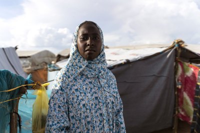 A woman with a matching dress and head scarf in a blue-and-white floral print stares at the camera. She stands in a refugee camp. 