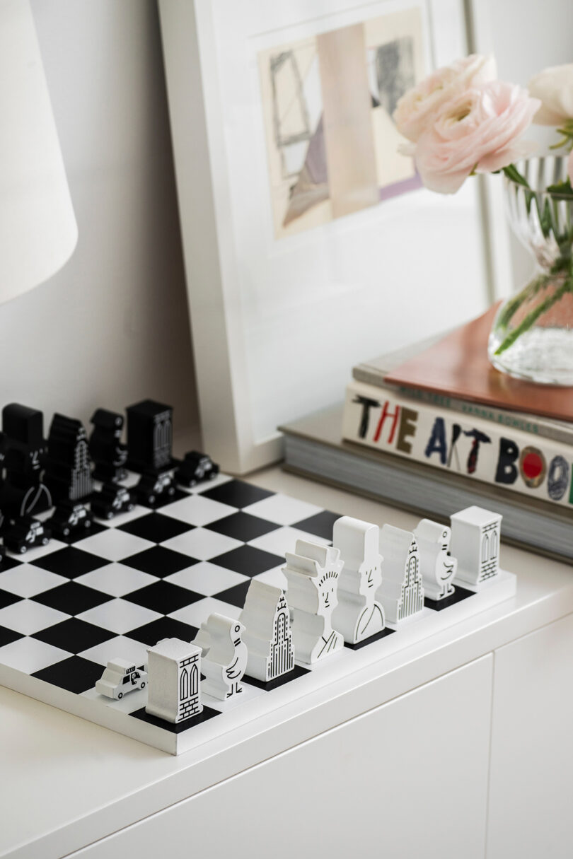 Stylized chess set on a white cabinet with artistic black and white pieces. Nearby is a stack of books, including 