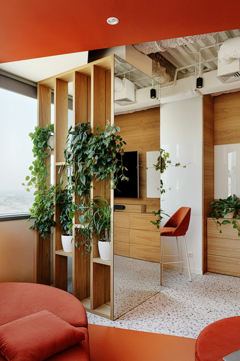 Modern office space with orange accents, a partition with potted green plants, a high chair, and a TV on a wooden cabinet. White and wood textures dominate the room.