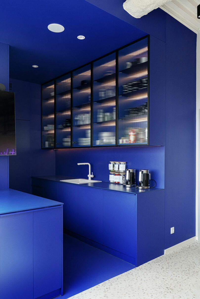 Modern kitchen with bold blue cabinetry, open shelves with various items, white countertop, and sleek appliances including a coffee maker and toaster.