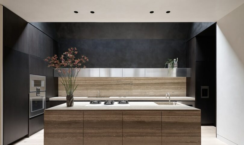Modern kitchen with dark wood cabinets, a marble island with a built-in cooktop, and metal shelves. A vase with tall flowers is on the island, and a plant is on the right shelf.