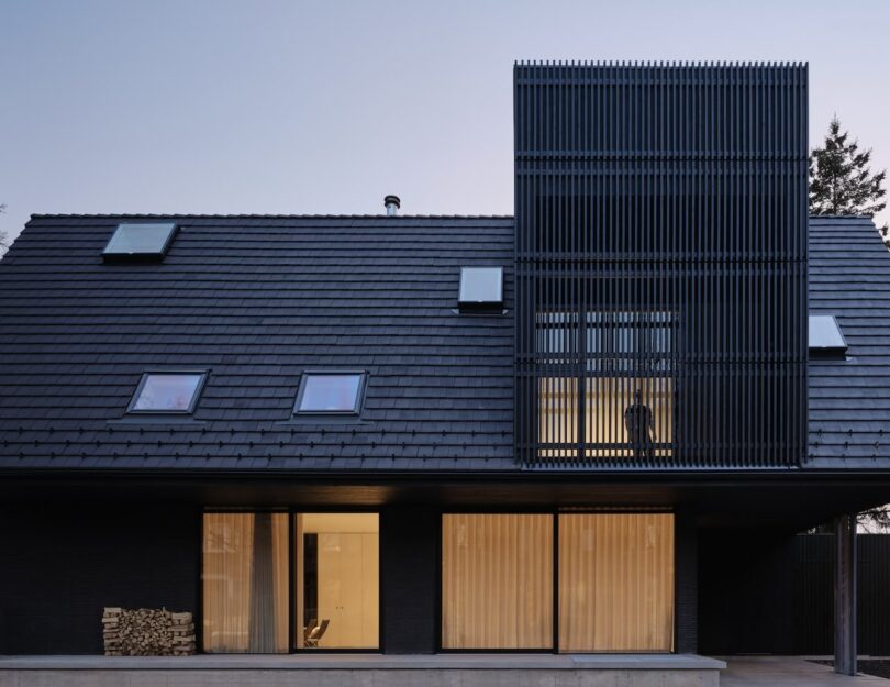 Modern house with a sloped roof featuring skylights and a section of vertical black slats. Large windows on the ground floor reveal illuminated interior spaces.