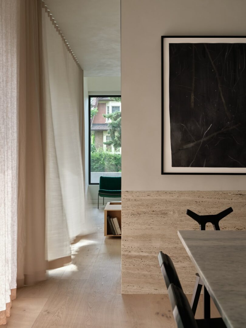 Modern interior with a marble table, a black chair, a framed abstract painting, beige curtains, and a window showing greenery outdoors.