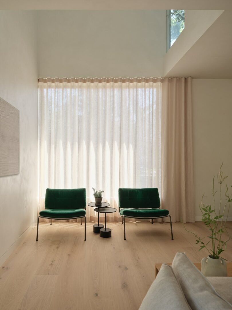 Two green chairs and a small round table with a plant are arranged in front of sheer curtains in a bright, minimalist room with light wooden flooring and a sofa edge visible.