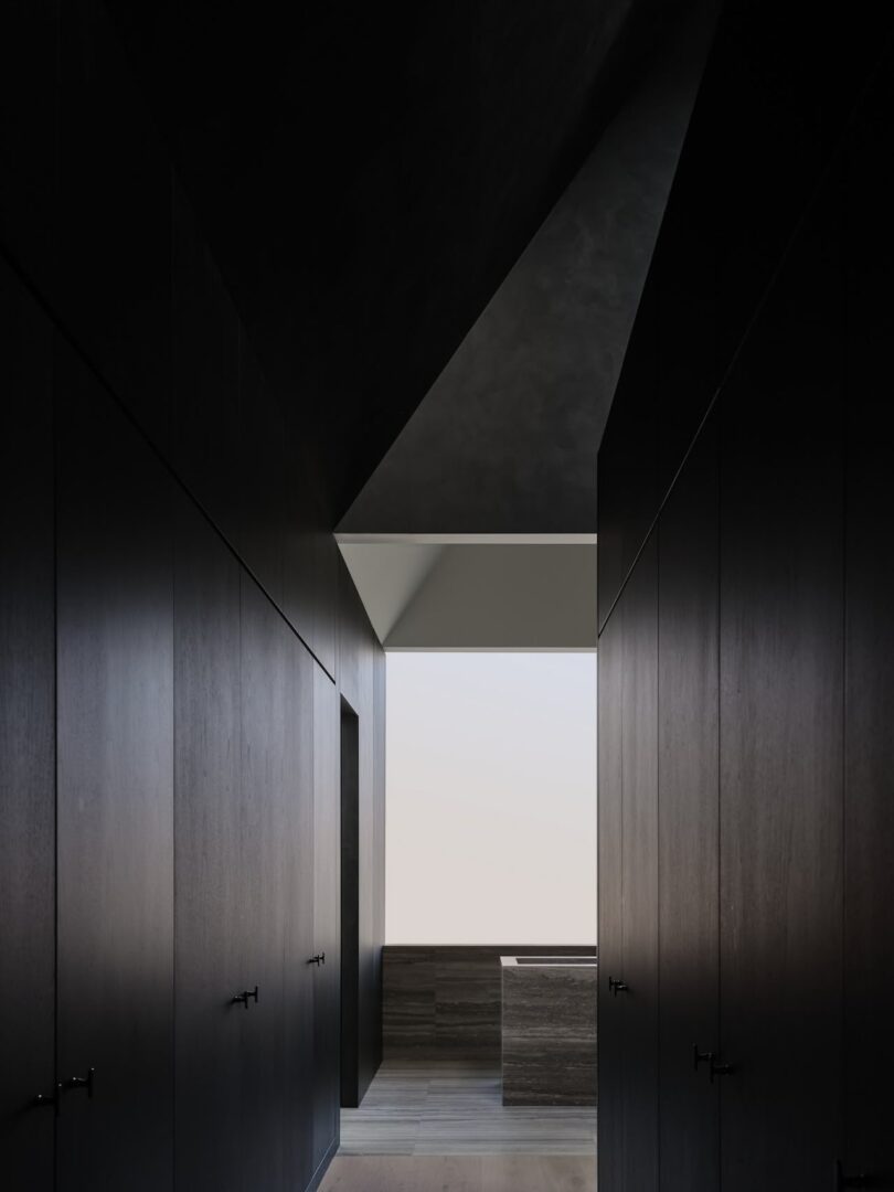 A dark hallway with modern, smooth walls leads to a bright, minimalist room with a large window and wooden flooring.