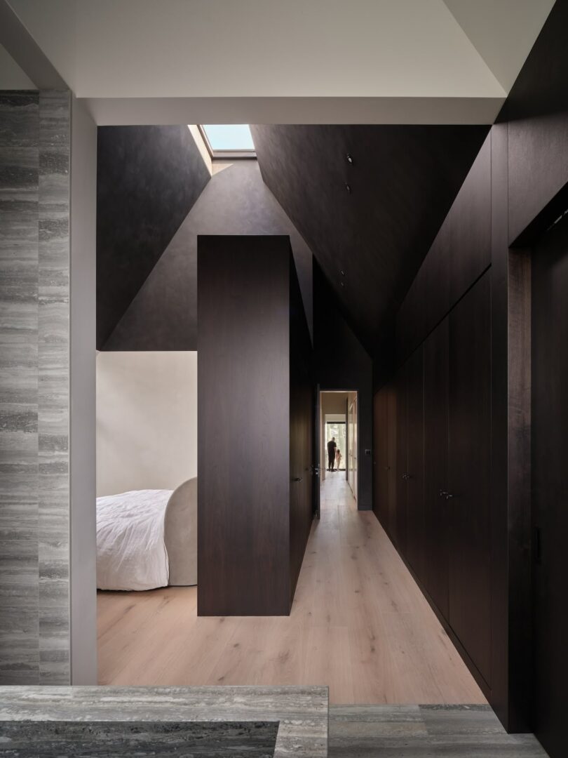 A modern bedroom features a dark wood hallway with high ceilings and a skylight. On the left, a bed with white bedding is visible, and a doorway at the end shows a distant figure.