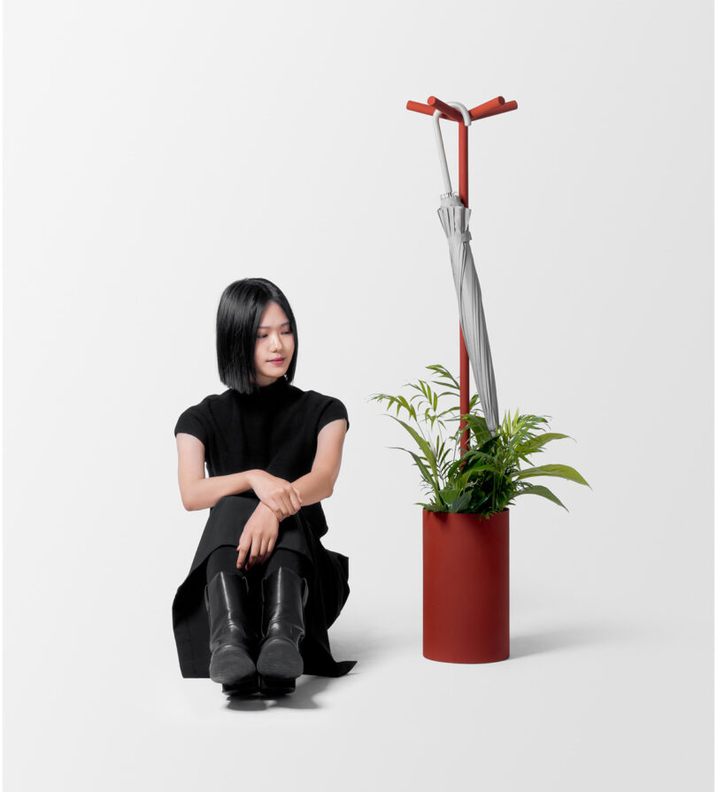 A woman in black clothing sits on the floor next to a red planter with a plant and a gray umbrella, isolated on a white background.