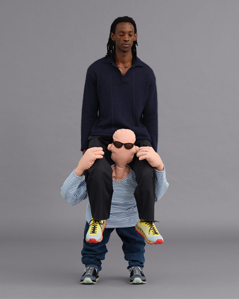 A person wearing a large puppet suit. The puppet is wearing sunglasses, striped shirt, jeans, and sneakers, against a gray background.
