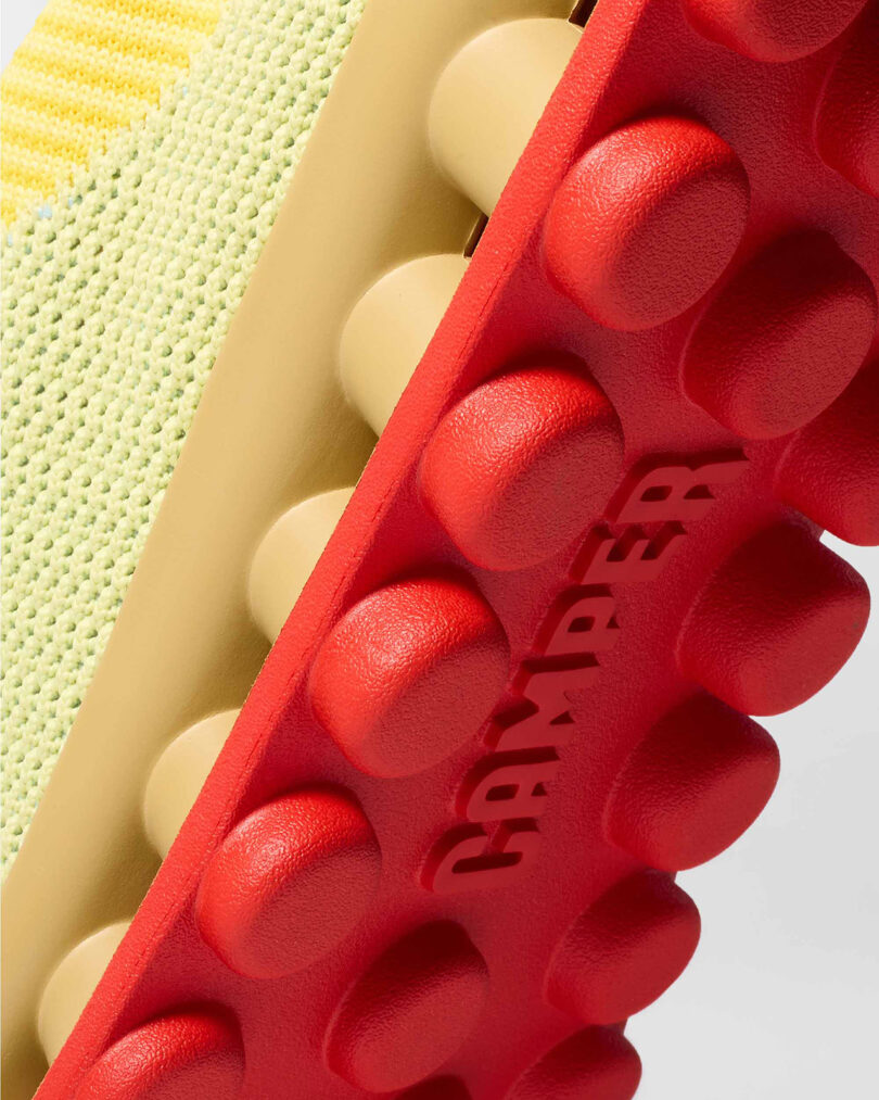 Close-up of a sneaker showing its textured yellow upper and red rubber outsole with round protrusions. The word 