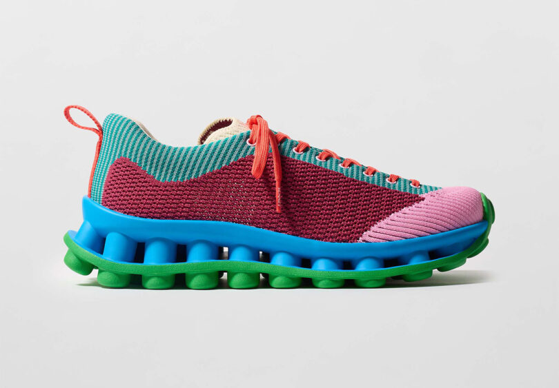 A colorful shoe featuring a pink toe, red mesh upper, turquoise laces, and a blue and green segmented sole, set against a plain background.